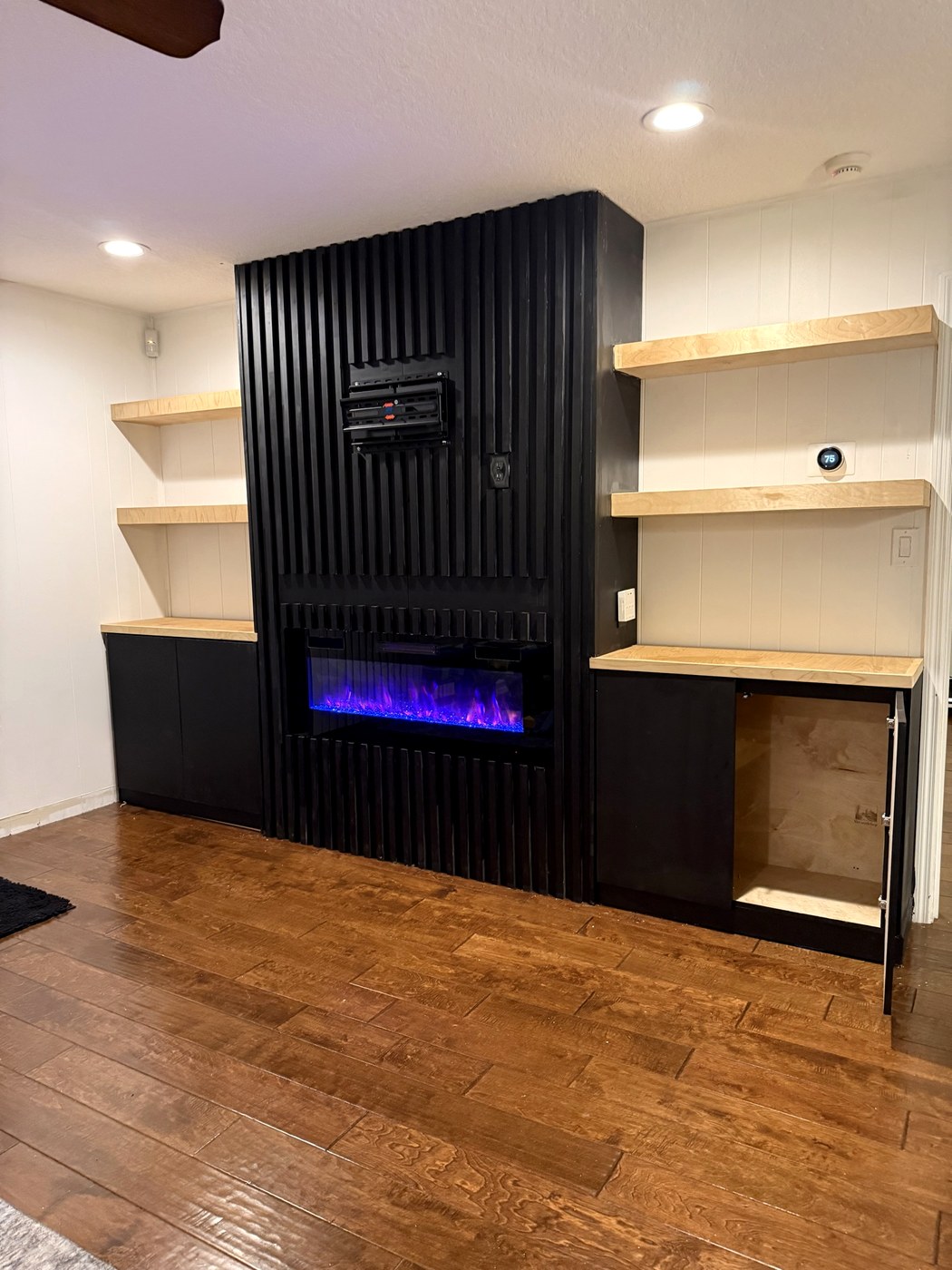Custom living room built-in: matte black fluted slat wall with recessed linear fireplace, maple floating shelves and black base cabinets with plywood interiors.