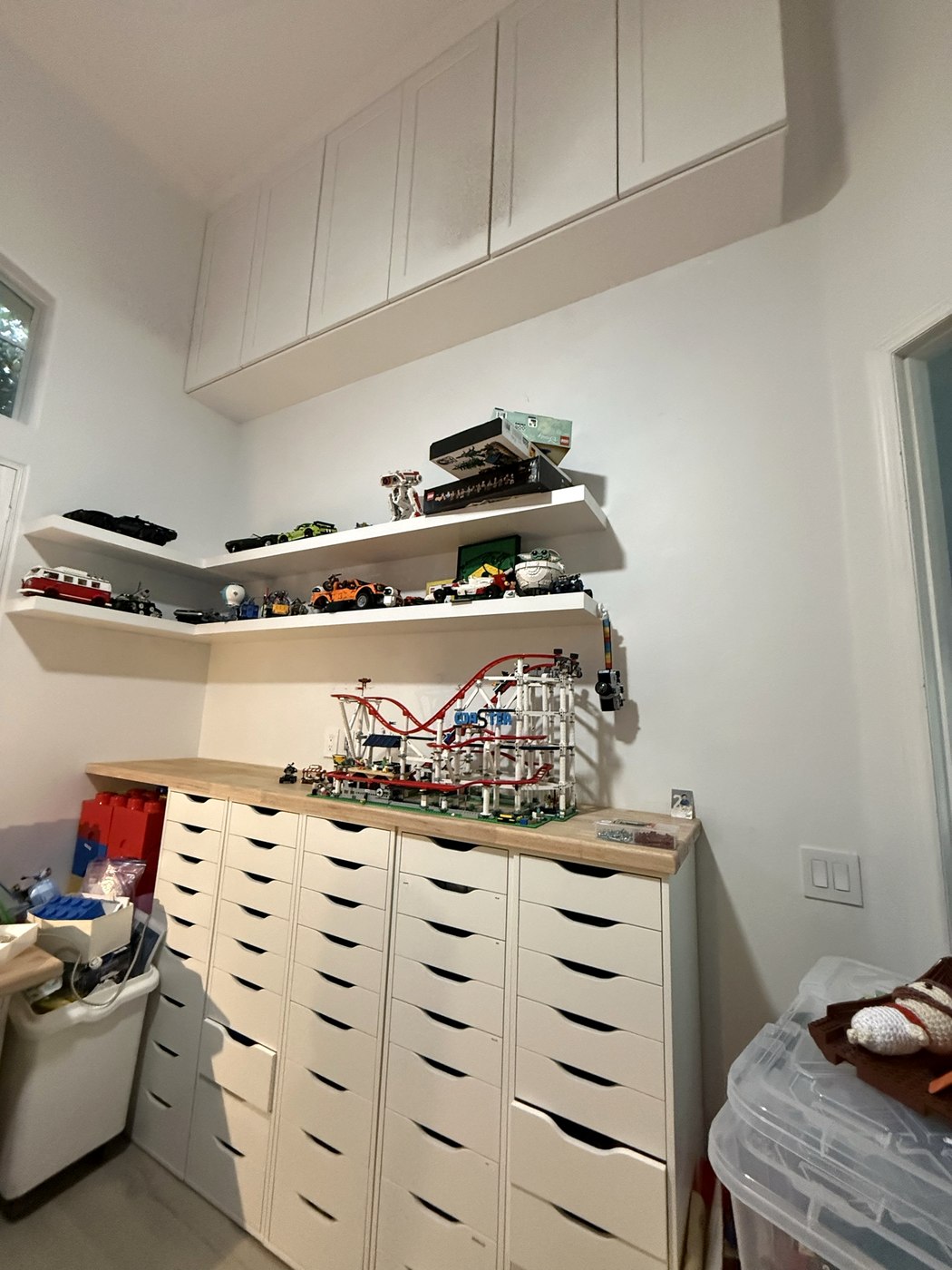 Hobby room wall with painted shaker upper cabinets running to the ceiling, white floating corner shelves and a butcher-block counter over banks of drawers.