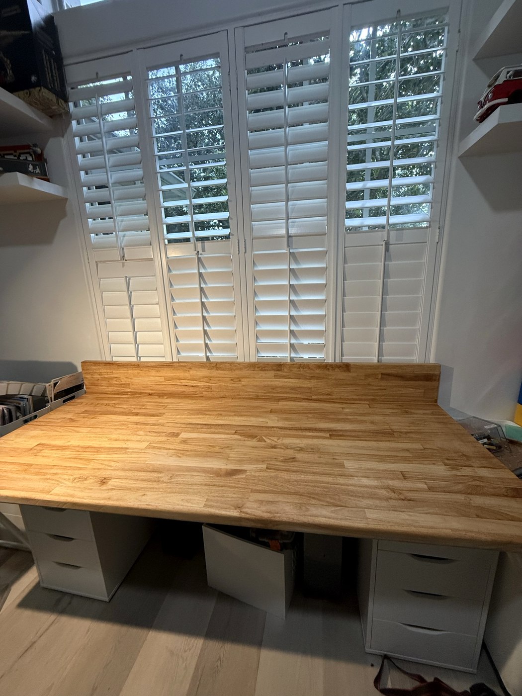 Butcher-block work desk built under plantation shutters, with a back lip and white drawer pedestals underneath.