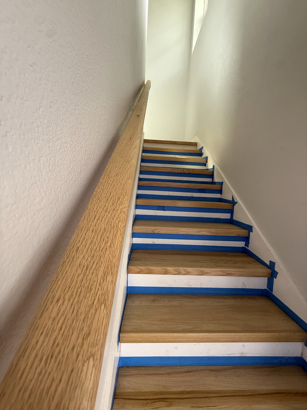 Staircase with new hardwood treads and a wall-mounted hardwood handrail, risers masked with blue tape mid-installation.